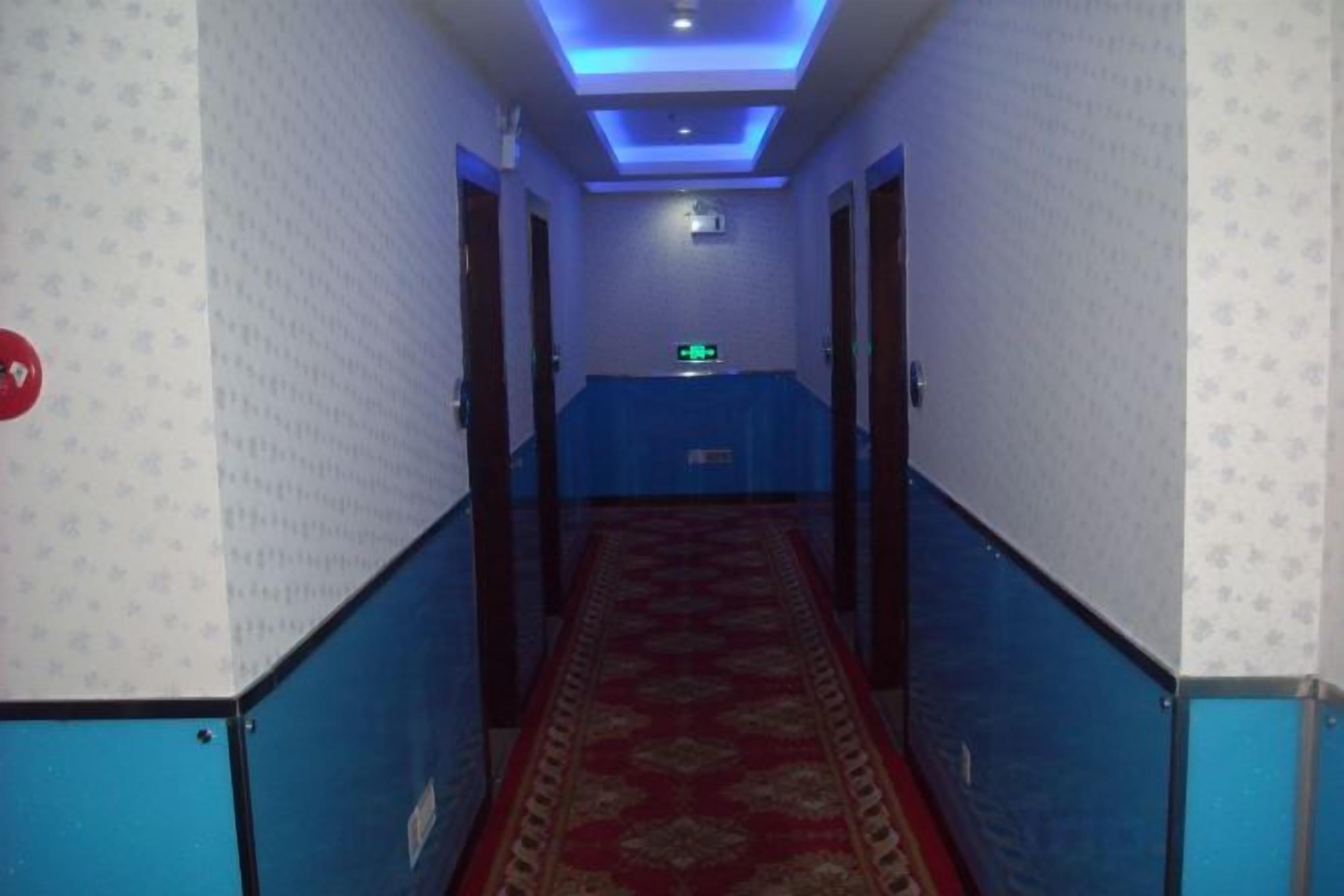 Corredor de hotel com parede estampada, carpete azul e um balcão de recepção no final.