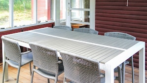 A covered outdoor dining area with a table and chairs, a door, and a view of greenery. - 6 Person Holiday Home in Blokhus (Blokhus)