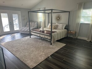 Une chambre à coucher avec un lit à baldaquin, un fauteuil gris, une table de chevet et une grande fenêtre avec des rideaux.