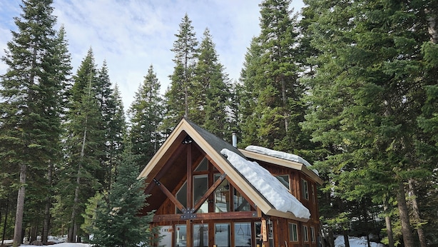 A wooden cabin with a steep roof, surrounded by tall evergreen trees and covered in snow.