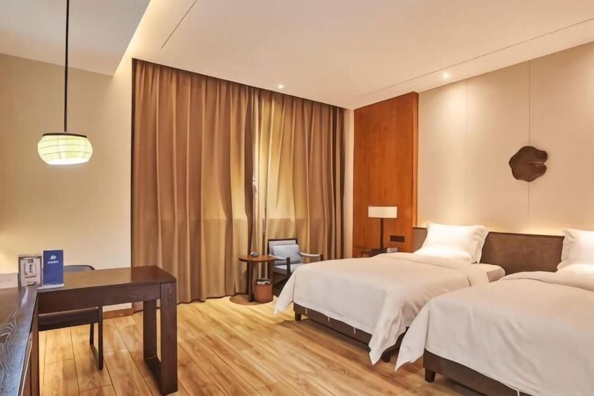 Ein Hotelzimmer mit zwei Betten, einem Schreibtisch, einem Stuhl, einer Lampe und einem großen Fenster mit Vorhängen.