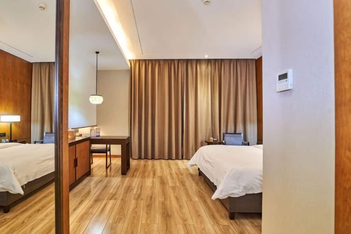 Ein Hotelzimmer mit zwei Betten, einem Schreibtisch, einem Stuhl, einer Lampe und einem großen Fenster mit Vorhängen.