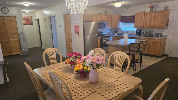 A dining area with a table set for four, surrounded by wooden chairs. A chandelier hangs above, and a vase with flowers is on the table.
