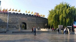 A stone wall with flags, a large tree, and people in a public square. - Qufu Ancient City Hotel (Sankong Scenic Area) (Qufu)