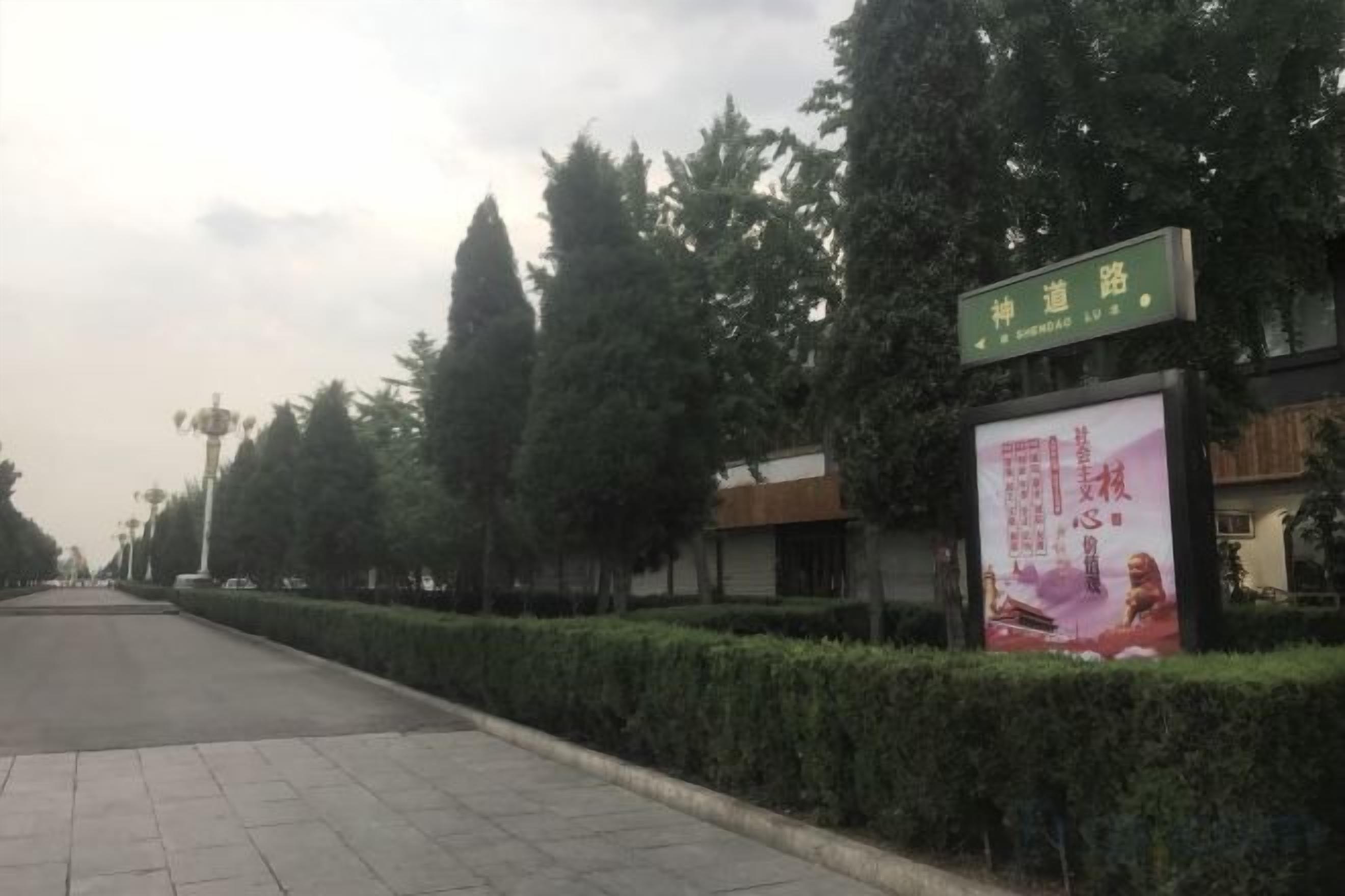 Un sendero pavimentado bordeado por setos cuidados y árboles altos. Un letrero verde con texto blanco está montado en un edificio a la derecha.