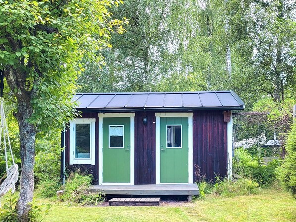 A small wooden cabin with two green doors and a metal roof, surrounded by lush greenery. - 4 Star Holiday Home in Frandefors (Frändefors)