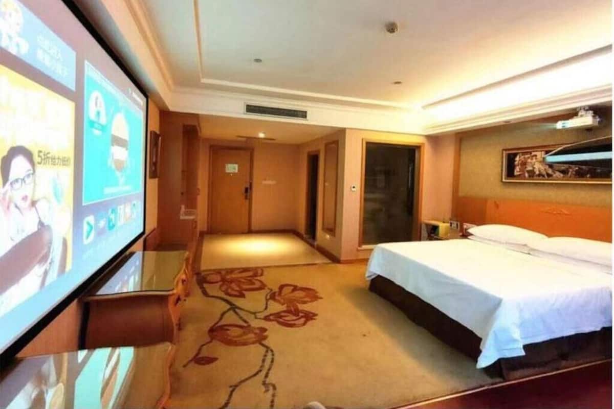 A hotel room with a bed, a TV screen, a carpet with a floral pattern, and a door leading to another room.