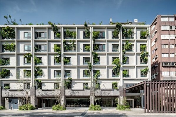 A modern multi-story building with a grid-like facade, balconies with greenery, and a covered entrance area.