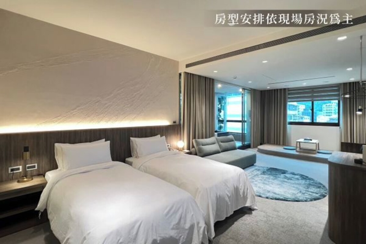 A modern hotel room with two beds, a seating area, and a large window.
