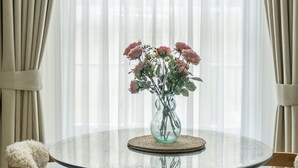 A dining area with a round glass table, beige curtains, and a vase with pink roses.