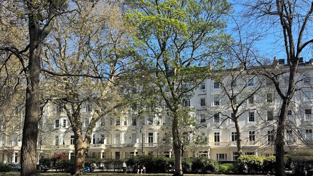 A park with mature trees and a row of white buildings in the background.