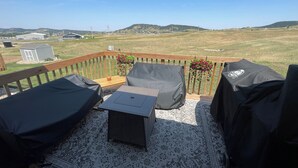 A deck with outdoor furniture, a small table, and a view of a grassy field and hills in the distance. - Sturgis Rally Rental! (Sturgis)