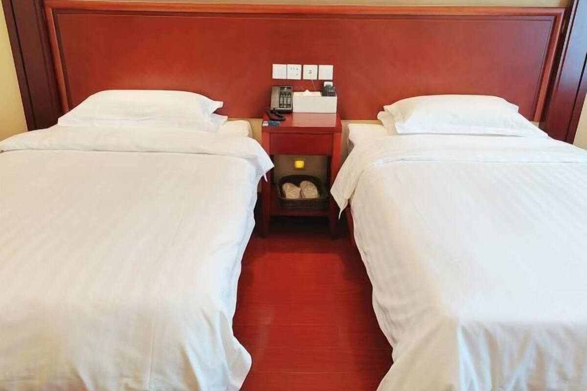 A hotel room with two single beds, a wooden headboard, a nightstand with a phone and a lamp, and a small storage space under the bed.