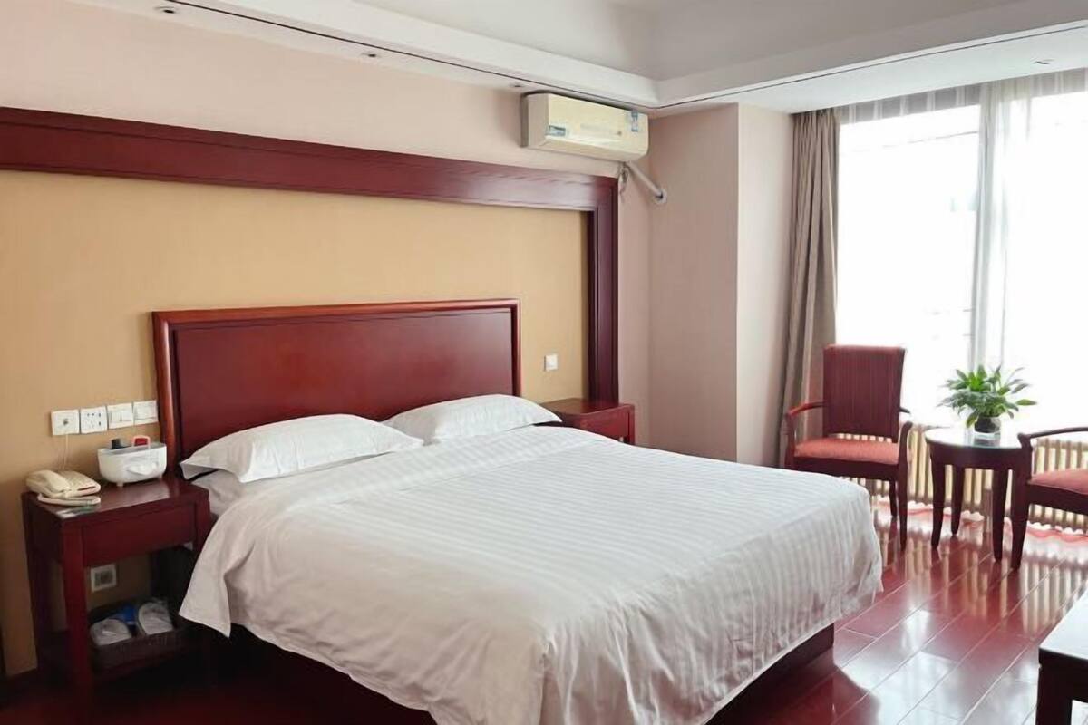 A hotel room with a large bed, a wooden headboard, a bedside table with a phone, a small table with a plant, and a chair.