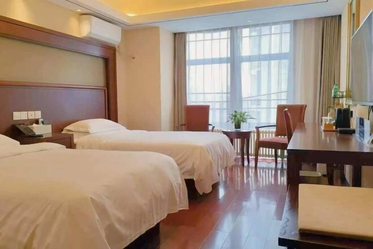 A hotel room with two beds, a wooden headboard, a large window with curtains, a small table with a chair, and a TV.