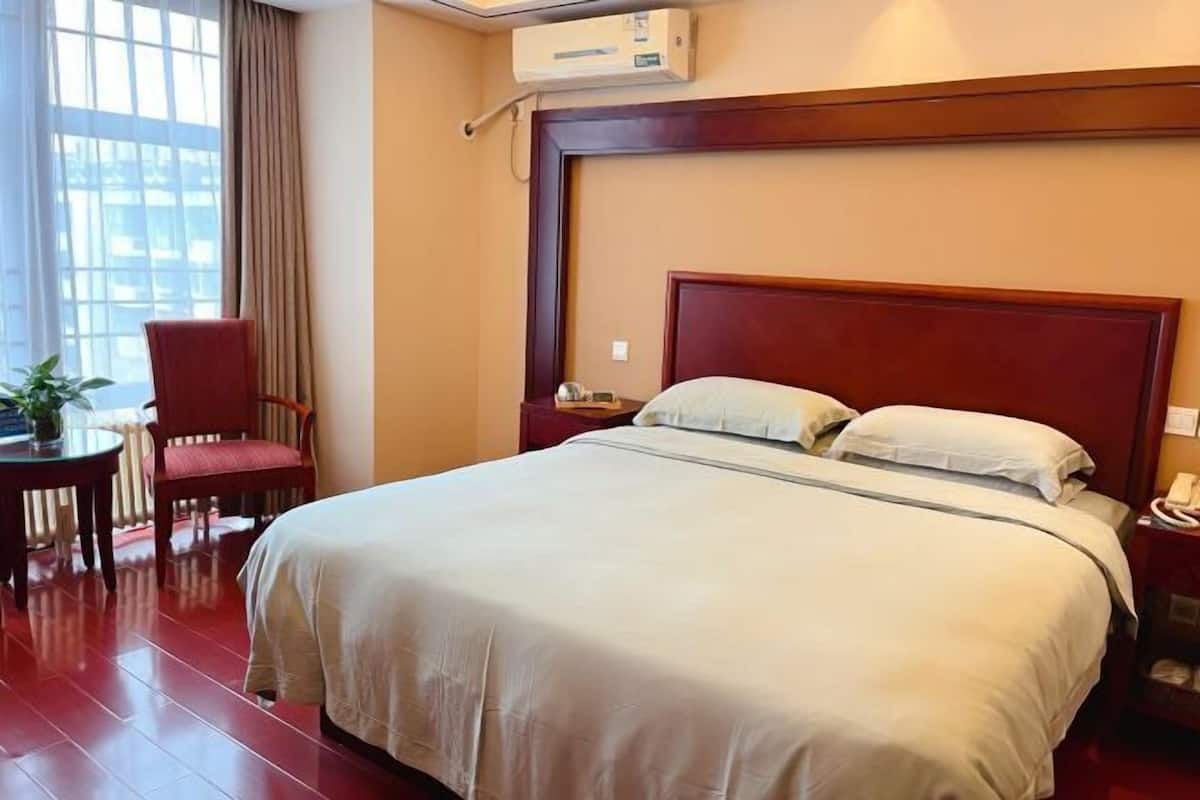A hotel room with a large bed, a desk with a chair, a bedside table, and an air conditioning unit.