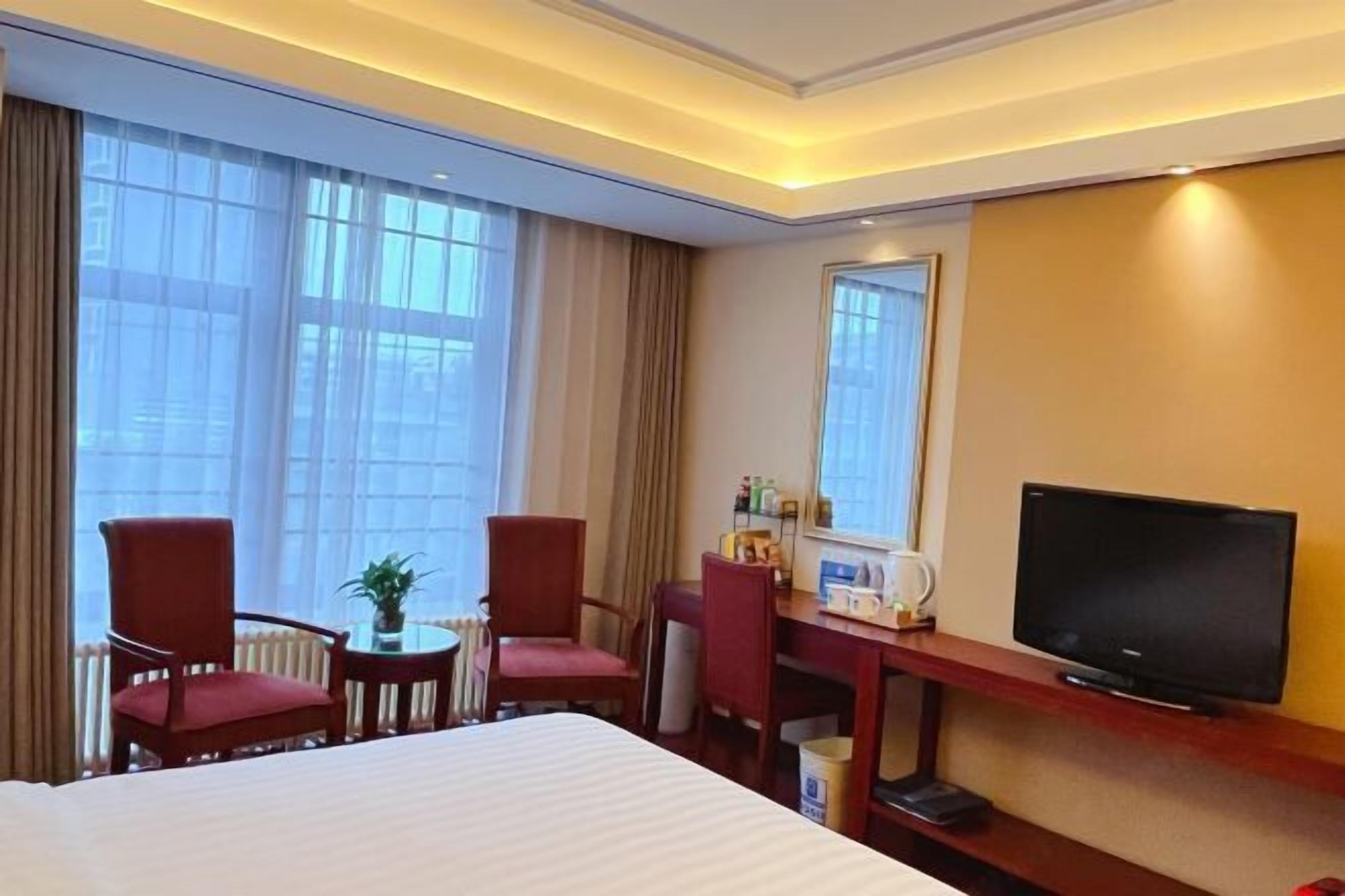 Quarto de hotel com uma cama grande, duas cadeiras vermelhas, uma mesinha com uma planta, uma televisão sobre um suporte de madeira e uma janela com cortinas.