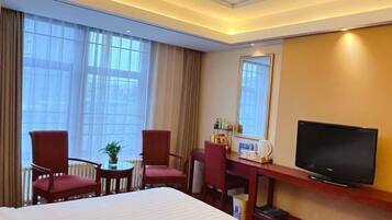 Quarto de hotel com uma cama grande, duas cadeiras vermelhas, uma mesinha com uma planta, uma televisão sobre um suporte de madeira e uma janela com cortinas.