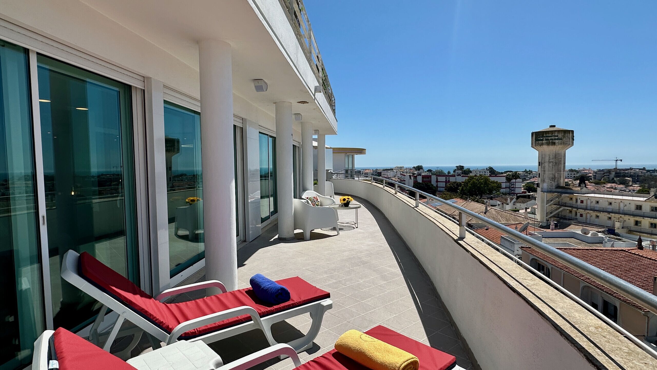 A balcony with sun loungers, a small table with a vase, and a view of the cityscape and a tower.
