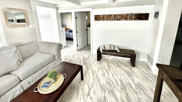 Un salon avec un canapé à rayures, un banc en bois et une table basse sur laquelle se trouve un plateau. Une pancarte sur le mur indique : « AU BORD DE LA MER ».