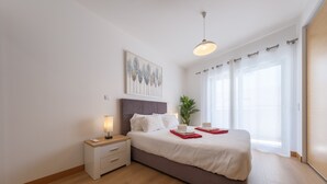 Ein modernes Schlafzimmer mit Bett, Nachttisch, Lampe und einem großen Fenster mit Vorhängen.