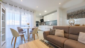 Ein modernes Wohnzimmer mit einem beige farbenen Sofa, einem runden Esstisch und zwei Stühlen. Im Bereich der Küche befinden sich weiße Schränke und ein weiterer Esstisch.