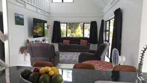 A spacious living room with a sofa, chairs, a dining table with fruit, and a TV on the wall.