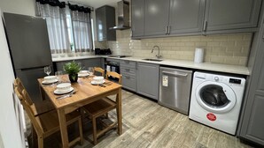 Una cocina moderna con armarios grises, una mesa de comedor de madera para cuatro personas, lavadora integrada y una ventana con cortinas.
