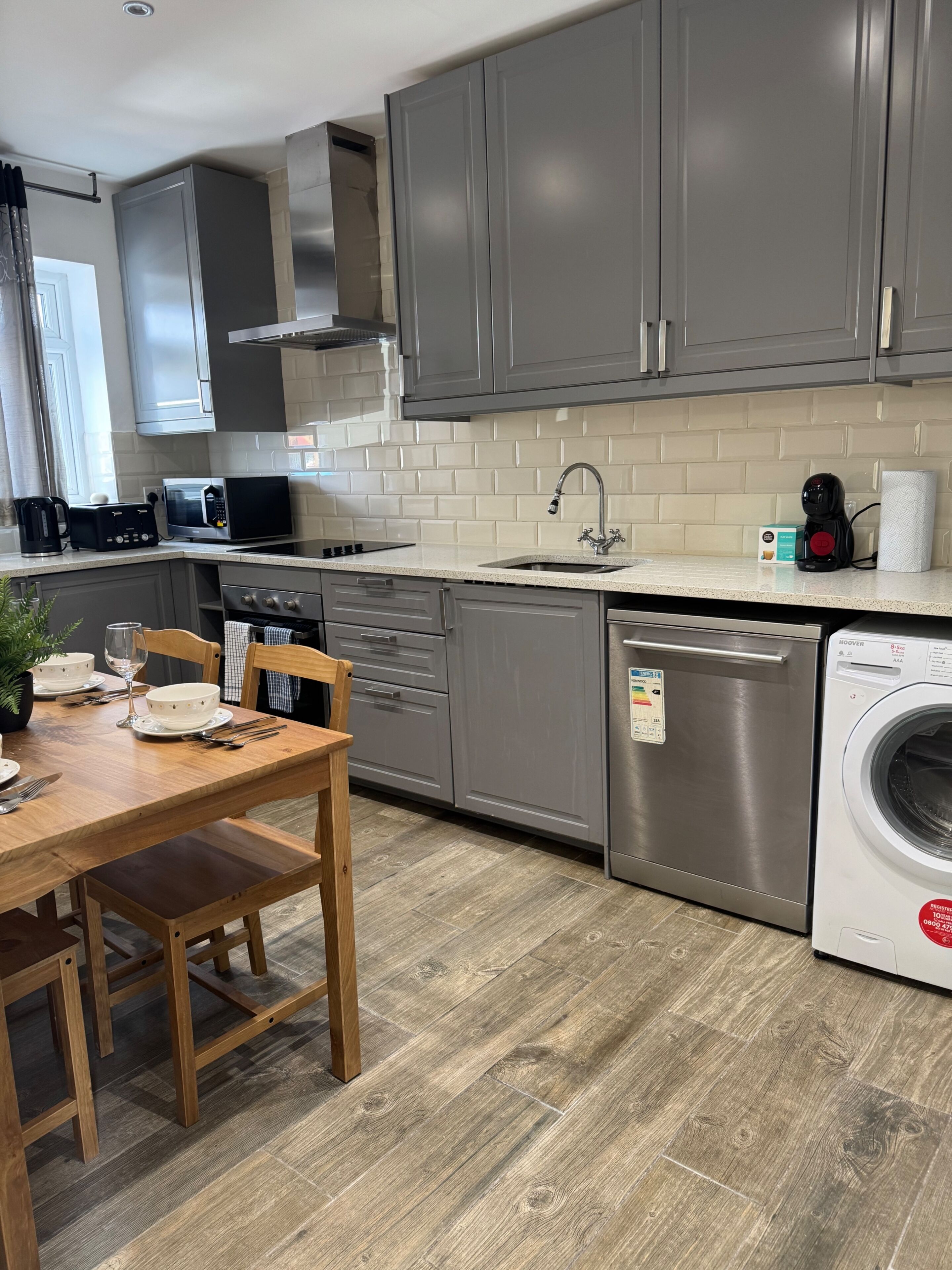 A modern kitchen with grey cabinets, a stainless steel range hood, a microwave, a sink, and a built-in washing machine.