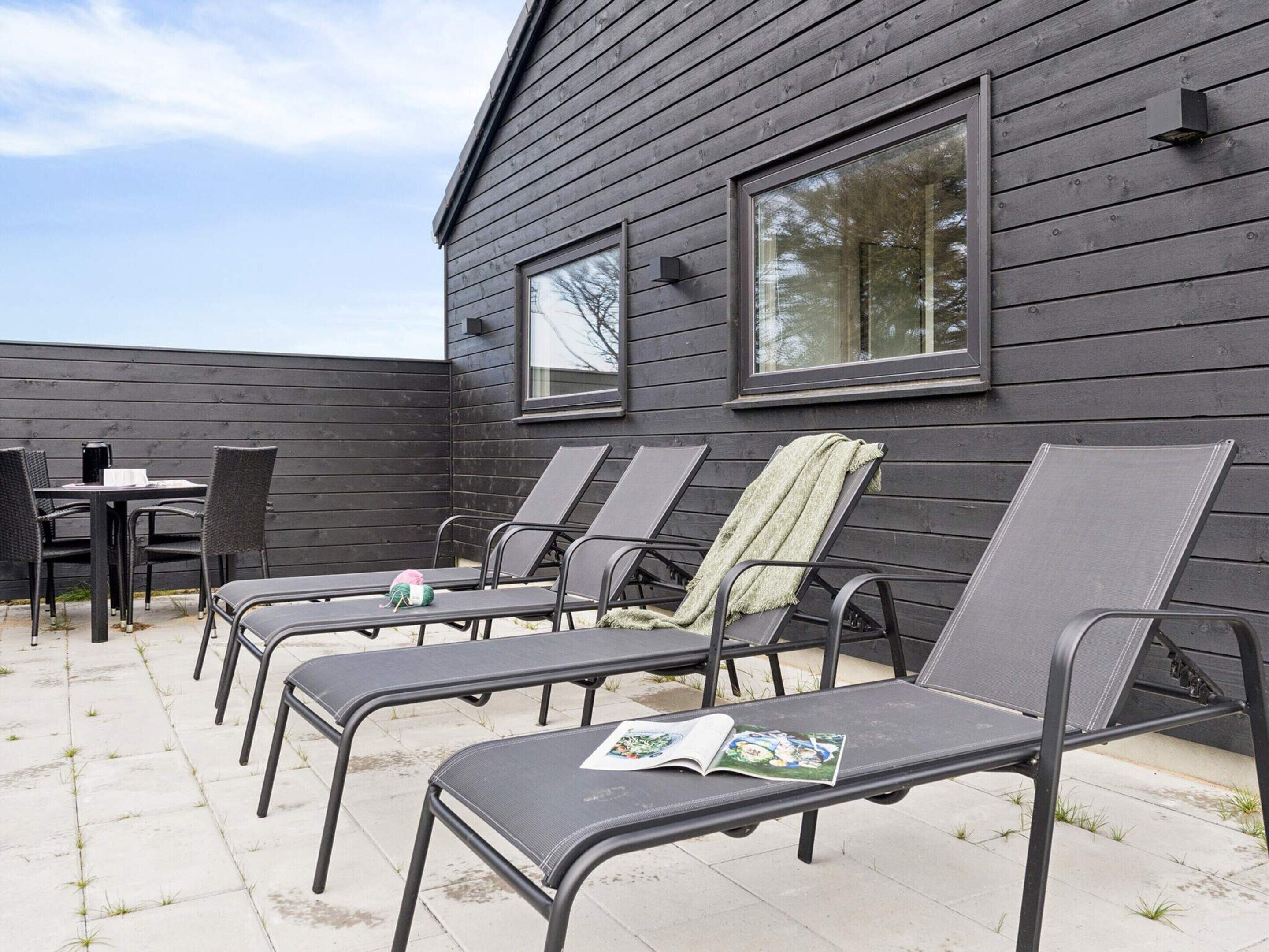 A patio with six lounge chairs and a table, a wooden building with large windows, and a clear sky.