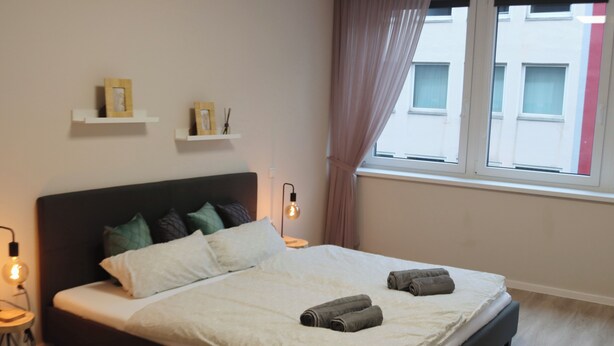 A neatly made bed with pillows and towels, a bedside lamp, and a shelf with decorative items. - Apartment in the heart of Braunschweig (Braunschweig)
