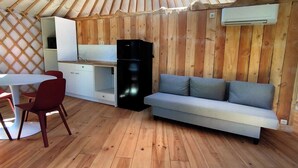 A compact, wooden interior with a sofa, a dining table with chairs, and a kitchenette area including a refrigerator and an air conditioning unit. - Camellia is a beautiful yurt 10 minutes walk from Pairi Daiza (Brugelette)