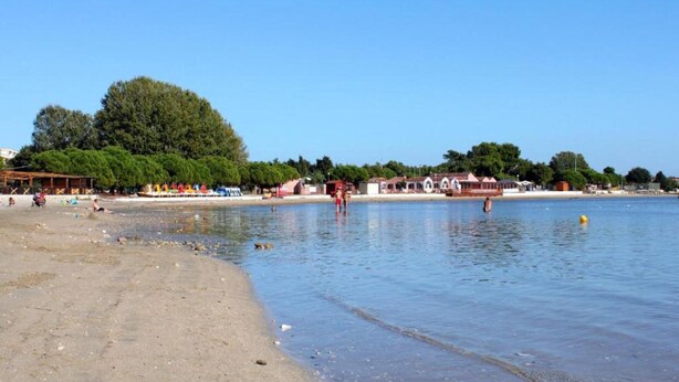 A beach with a sandy shore, a calm water body, and a row of buildings in the background. - Holiday Home With Garden, sea View (Medulin)