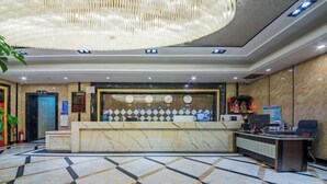Zona de recepción de un hotel con suelo de mármol, un mostrador de recepción, una gran lámpara de araña y un panel informativo digital.