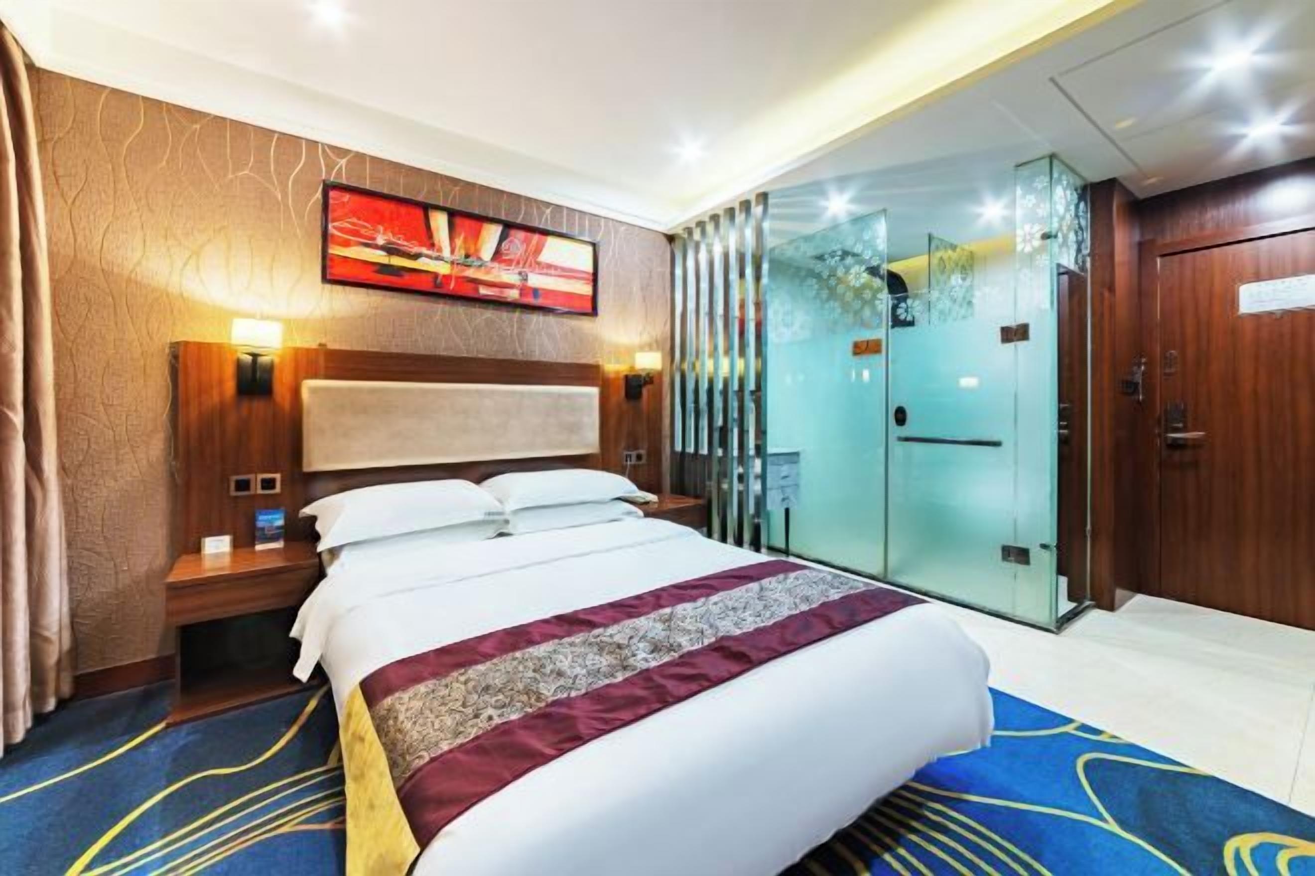 A hotel room with a large bed, a nightstand, a wall-mounted TV, and a glass door leading to a bathroom.