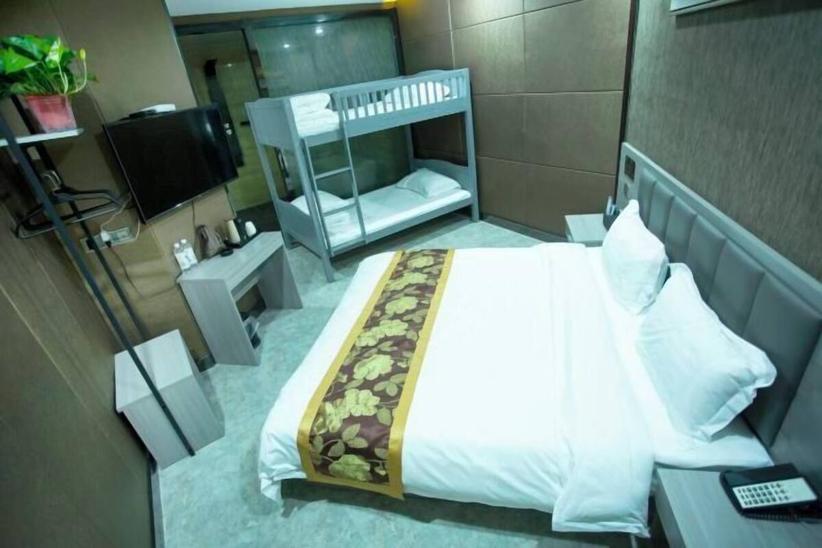 Hotelkamer met een stapelbed, een bed met bloemenprint, een televisie, een klein bureau met stoel en een kamerplant.
