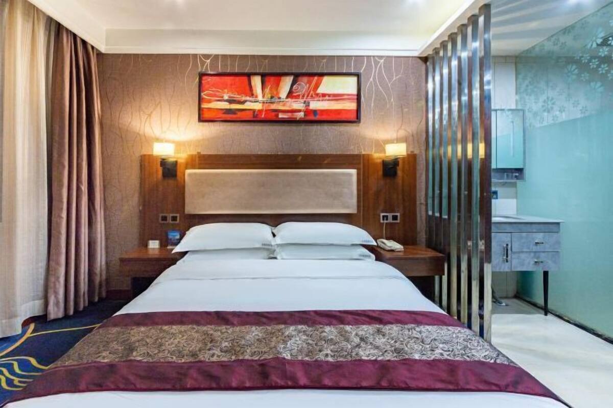 Een hotelkamer met een groot bed, hoofdbord, bedlampen, nachtkastje, spiegel en een raam met gordijnen.