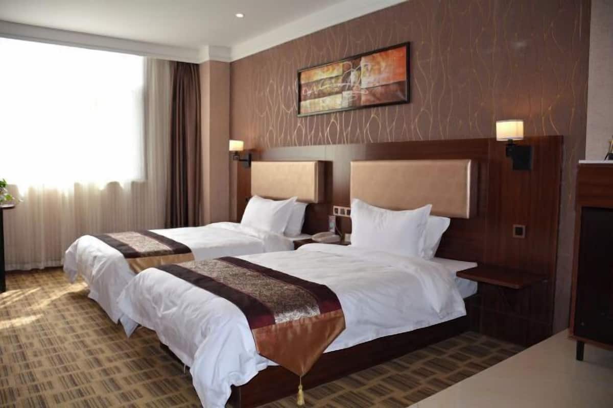 Een hotelkamer met twee bedden, een groot raam met gordijnen, een schilderij aan de muur en een houten hoofdbord.