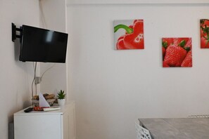 A room with a flat-screen TV mounted on the wall, a small cabinet with snacks, a potted plant, and a dining table with a chair.