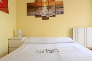 A hotel room with a bed, a nightstand with a lamp, a wall with a sunset painting, and a radiator.