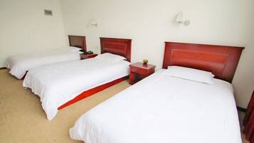 A hotel room with three single beds, each with a wooden headboard and white bedding. There are bedside tables with lamps and a small table between the beds.