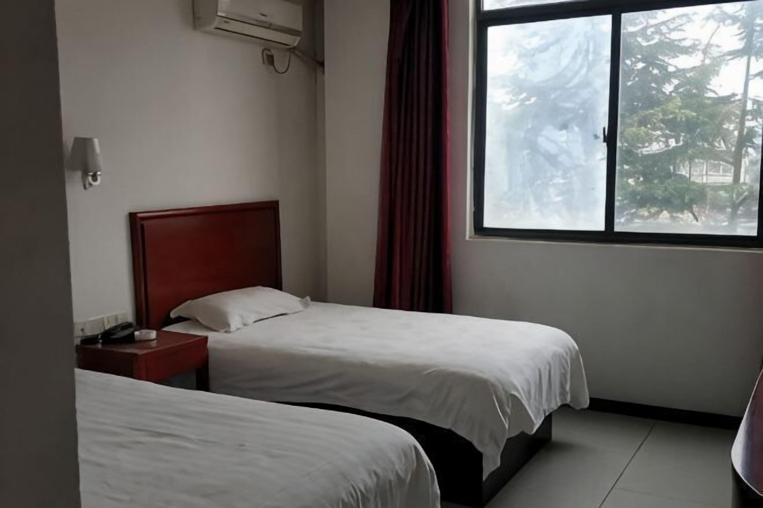Una camera d'albergo con due letti, una testiera rossa, un comodino, una finestra con vista sugli alberi e un condizionatore d'aria.