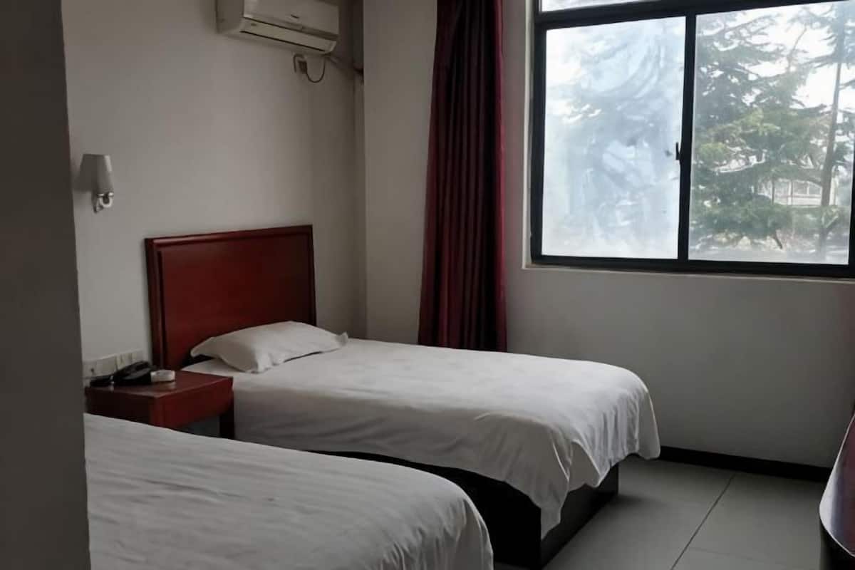 A hotel room with two beds, a red headboard, a nightstand, a window with a view of trees, and an air conditioning unit.
