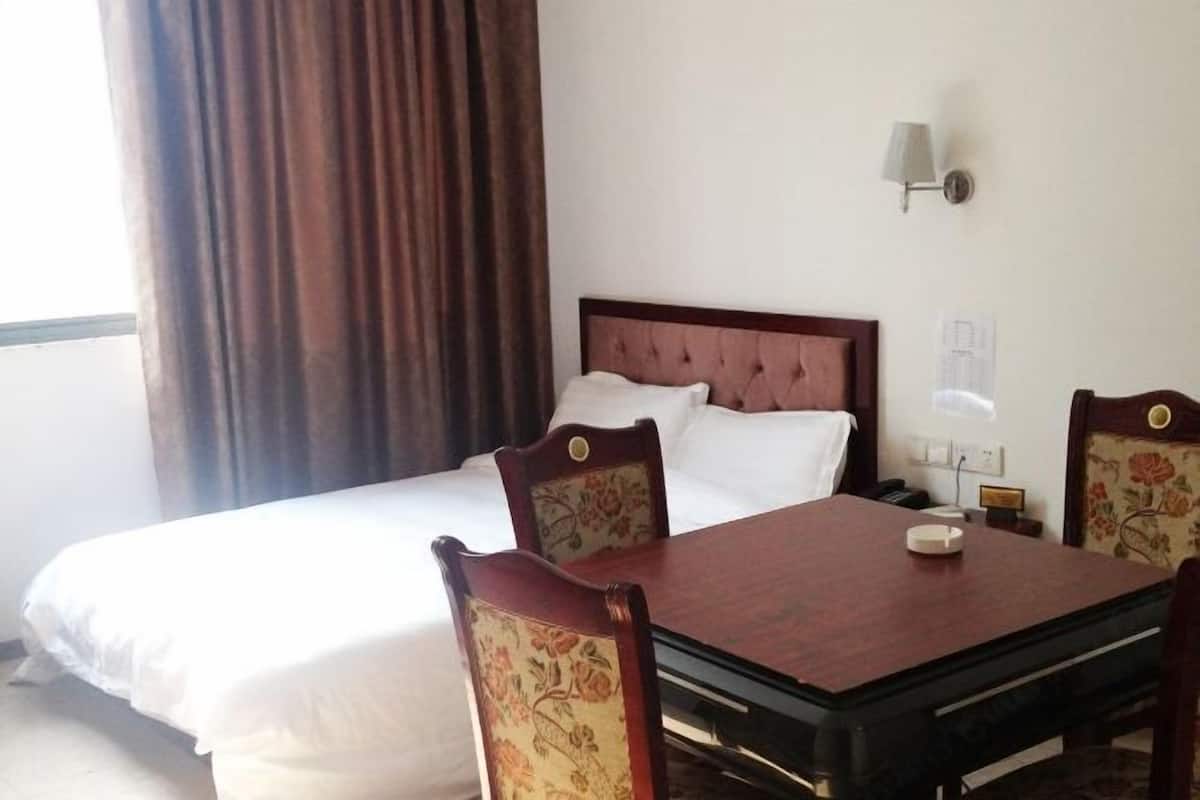 A hotel room with a bed, a wooden table, two chairs, a lamp, and a window with curtains.