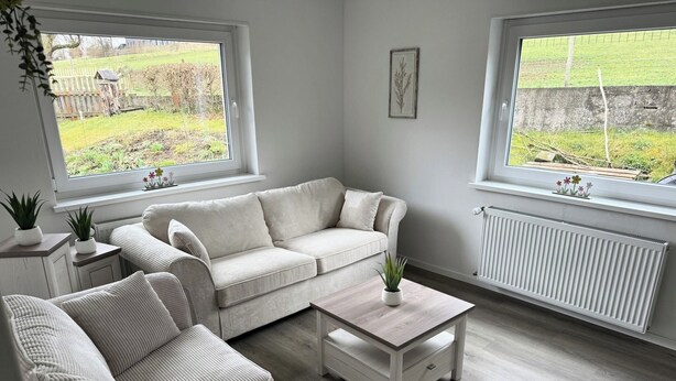 Uma sala de estar moderna com um sofá bege, uma mesa de centro e duas janelas com vista para um jardim.