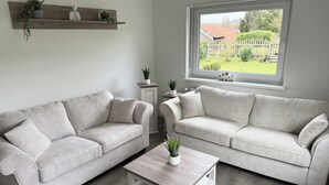 Uma sala de estar com dois sofás claros, uma mesa pequena branca e uma janela com vista para um jardim.