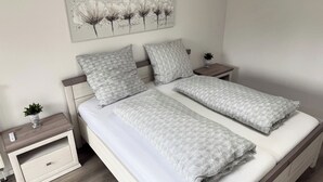 Uma cama de casal bem arrumada com um cobertor cinza acolchoado e almofadas, uma mesinha de cabeceira com uma planta e uma obra de arte na parede acima da cama.
