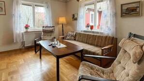 Ett mysigt vardagsrum med trägolv, en soffa, en fåtölj, ett bord och två fönster med gardiner.
