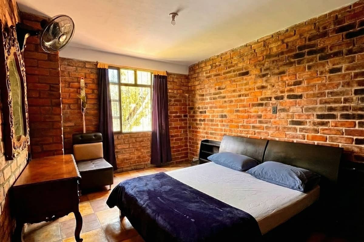Un dormitorio con pared de ladrillo, una cama con cabecera, una mesita pequeña, una silla y una ventana con cortinas.