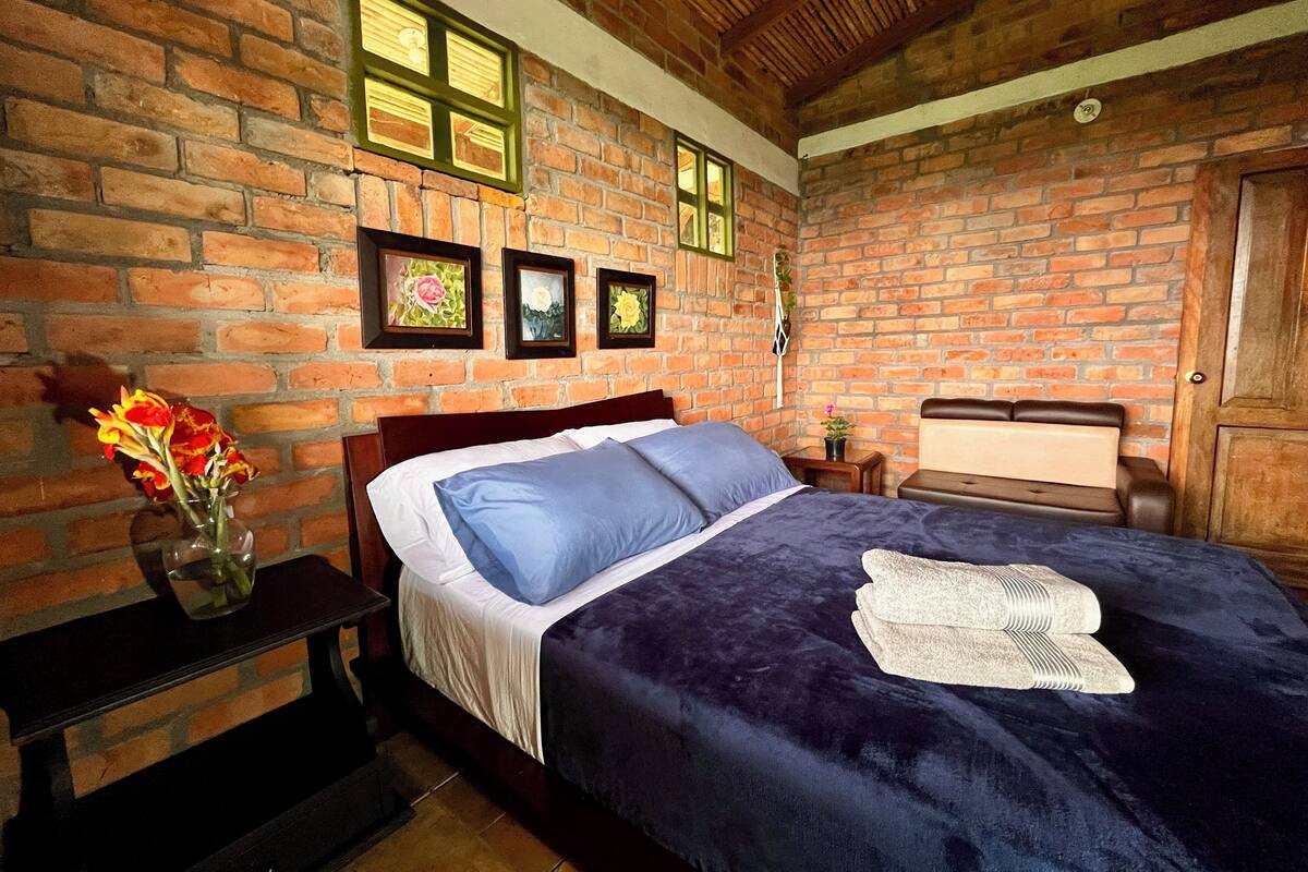 Un dormitorio con pared de ladrillo, una cama con ropa de cama azul, cabecera de madera y una mesita de noche con un florero.